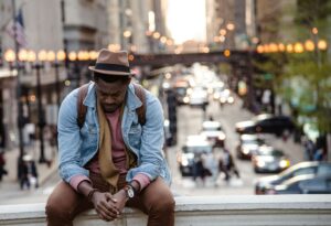 a man wearing a leather jacket, sitting on a ledge in a city, gazing down